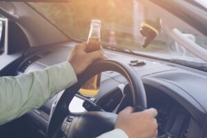 A person driving a car in a dress shirt holds a beer bottle in one hand.