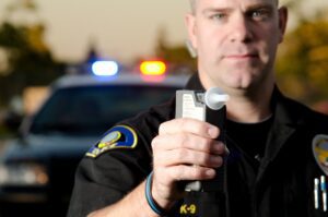 A police officer holds a Breathalyzer test with flashing lights on a police cruiser in the background.