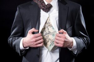 A man is holding open his suit and shirt to reveal $20 bills stuffed in it.