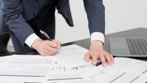 A man in a suit leaning over a desk bearing a number of documents and a laptop as he holds a pen.