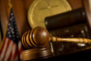 A close-up of a gavel in a court room with a flag and Judge’s chair in the background.