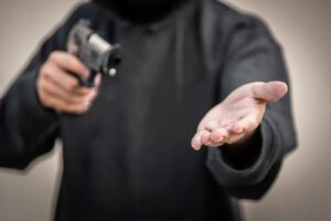 Person wearing dark hoodie pointing a gun and extending an open hand, appearing to demand something.