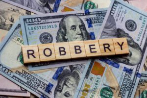 Wooden letter tiles spelling "ROBBERY" placed on top of scattered U.S. hundred-dollar bills.